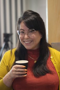 Angela Martinez Dy, a dark haired woman with gold wire rimmed glasses and a gold necklace, wearing a rust coloured top and mustard cardigan holding a paper cup of coffee.
