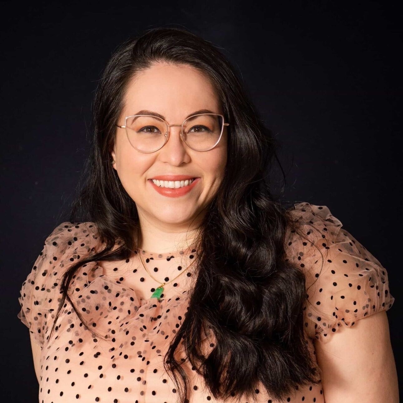 A smiling Filipina mestiza woman wearing gold glasses with chest length wavy brown hair in a pink short sleeved top with black polka dots, wearing a gold chain with a jade pendant.