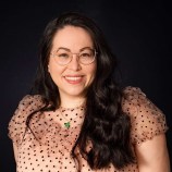 A smiling Filipina mestiza woman wearing gold glasses with chest length wavy brown hair in a pink short sleeved top with black polka dots, wearing a gold chain with a jade pendant.