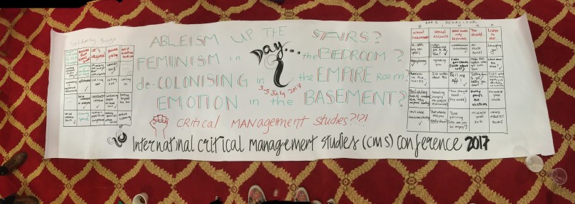 Long white paper banner on red and gold carpet reading: Ableism Up the Stairs? Feminism in the Bedroom? Decolonizing in the Empire Room? Emotion in the Basement? Critical Management Studies?! Photo: Angela Martinez Dy.
