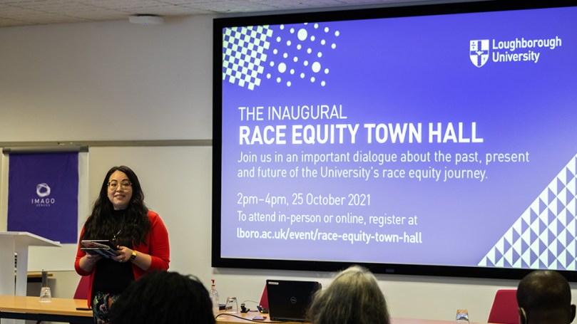 An Asian American woman in glasses and a red jacket stands in front of a purple screen reading:Loughborough University Inaugural Race Equity Town Hall, 25 October 2021